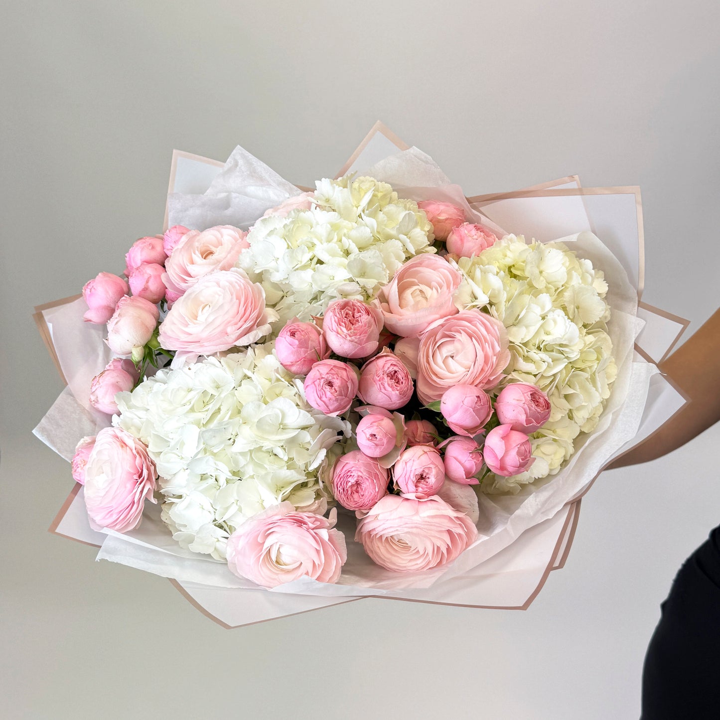 Luxusní kytice od Urban Bloom s bílými hortenziemi, růžovými ranunkuly a keřovými růžemi, zabalená v elegantním papíru.